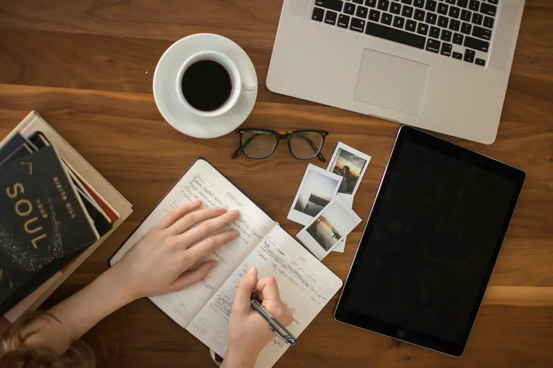 Someone writing in a notebook next to a laptop and a cup of coffee