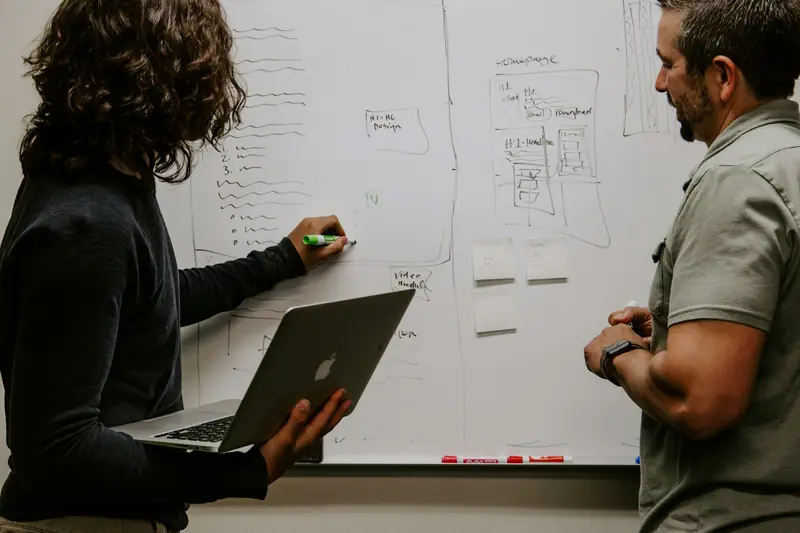 Two men talking at a whiteboard, one holding a laptop