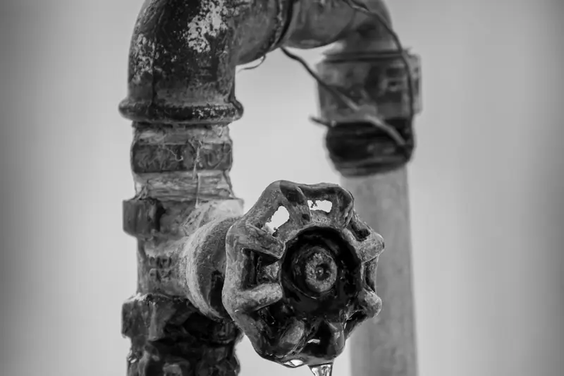 Een oude, roestige waterleiding met een kraan waar een druppel uit valt, in zwart-wit